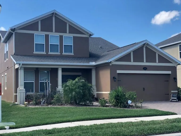 A cozy, two-story suburban home with a newly installed dark-gray asphalt shingle roof, light-colored exterior siding, and white-trimmed windows. The house is framed by lush green trees and is set beneath a partly cloudy blue sky—emphasizing the fresh, neat roofing work.