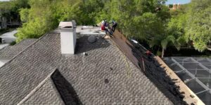Photo of a residential home with a dark gray shingle roof, where four workers are on the rooftop performing repairs, surrounded by trees and a neighboring house under a partly cloudy sky.
