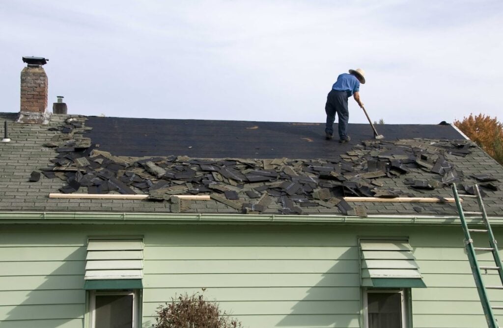 A man in a white hat, light blue shirt, and dark blue pants is scraping and removing old shingles from a house roof, illustrating the manual labor involved in roof replacement.